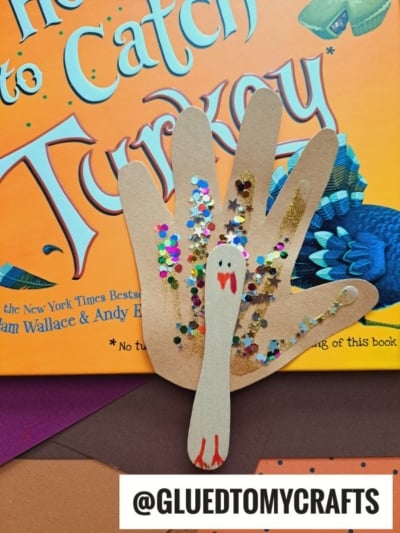 A handprint turkeys craft made from brown paper, decorated with colorful sequins and a drawn face, is held in front of the book How to Catch a Turkey. The handle @GLUEDTOMYCRAFTS is visible.