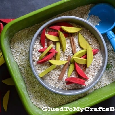 A green bin filled with oats, a round metal pan, a blue scoop, cinnamon sticks, and red and yellow wooden pieces resembling apple slices creates an inviting Apple Pie sensory bin.