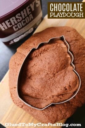 A mound of hot chocolate play dough with a leaf-shaped cookie cutter pressed into it. A container of Hershey’s cocoa sits nearby. Text on the image reads “CHOCOLATE PLAYDOUGH” and “GluedToMyCraftsBlog.com”.