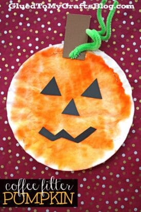 This Coffee Filter Pumpkin craft features a round pumpkin made from a coffee filter, colored orange with a green pipe cleaner stem, brown paper accents, and a smiling jack-o’-lantern face on a red polka dot background.