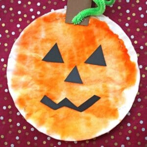 This Coffee Filter Pumpkin craft features a round pumpkin made from a coffee filter, colored orange with a green pipe cleaner stem, brown paper accents, and a smiling jack-o’-lantern face on a red polka dot background.