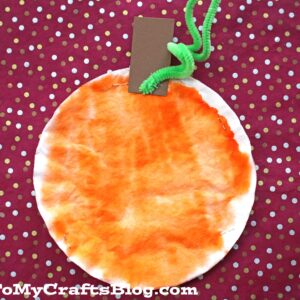 A round Coffee Filter Pumpkin, painted orange with a brown paper stem and green pipe cleaner leaf, sits on a red background with gold polka dots