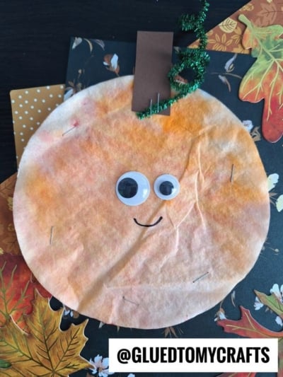 A Coffee Filter Pumpkin craft made from an orange-dyed coffee filter, with googly eyes, a drawn smile, a brown paper stem, and a green pipe cleaner vine, placed on a fall-themed background. @GLUEDTOMYCRAFTS is labeled at the bottom.