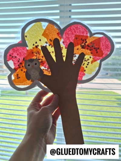A hand holds up a fall-themed craft shaped like a tree with a brown paper trunk and a tissue paper canopy, featuring star stickers and a small owl sticker—resembling Stained Glass Fall Trees—against a window with blinds.