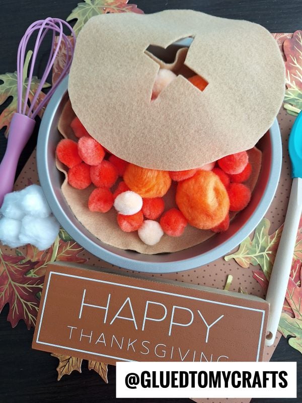 A felt pumpkin pie topped with pom-poms sits on a plate beside a whisk, chef’s spoon, and cotton balls. Fall leaves surround a sign that reads Happy Thanksgiving.