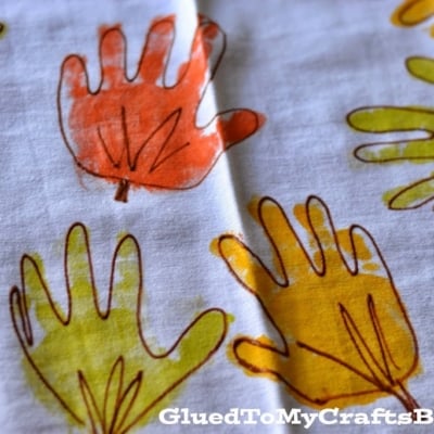 A white cloth decorated with orange and yellow handprint art, each outlined in brown to resemble autumn leaves, creates festive Handprint Leaf Napkins. The text GluedToMyCraftsBlog.com appears in the bottom right corner.