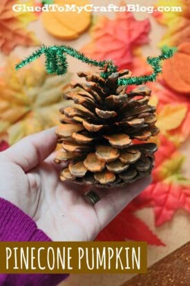 A hand holds a Pinecone Pumpkin—decorated with orange paint and green pipe cleaners to resemble a pumpkin. Colorful felt leaves are scattered in the background. Text reads PINECONE PUMPKIN.