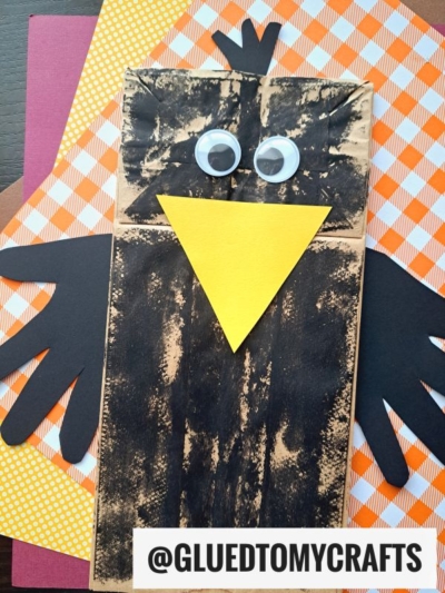 A Paper Bag Crow craft with big googly eyes, a yellow beak, and black paper wings sits on orange and white gingham paper. The handle @GLUEDTOMYCRAFTS is visible.