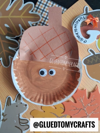 A charming Paper Plate Acorn craft, featuring googly eyes and a coat of brown paint, is surrounded by vibrant paper leaves, an adorable fox, and a round sticker labeled @GLUEDTOMYCRAFTS, all set against a cozy fall-themed background.