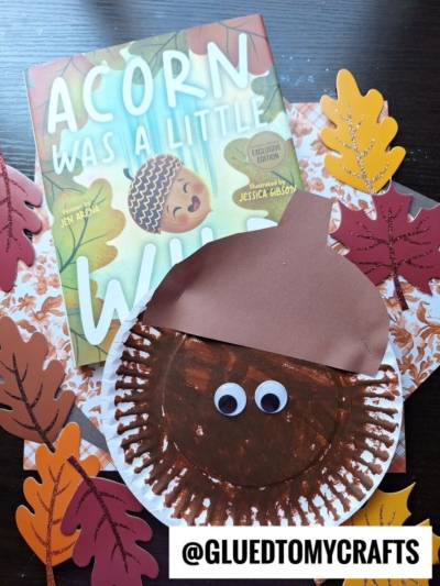 A children’s book titled “Acorn Was A Little Wild” is surrounded by colorful paper leaves and a whimsical Paper Plate Acorn craft with googly eyes. The handle @GLUEDTOMYCRAFTS is shown at the bottom.