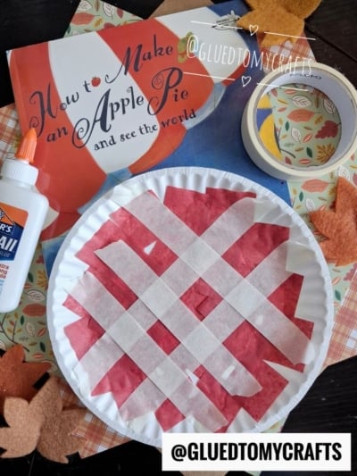 A children’s craft project: a paper plate cherry pie with red paper and white tape arranged like a lattice crust, surrounded by fall leaves, glue, tape, and the book How to Make an Apple Pie and See the World.