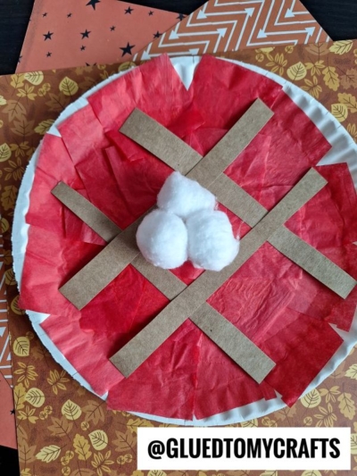 A delightful paper plate cherry pie craft, this project features red tissue paper filling, brown paper strips for the crust, and three fluffy cotton balls on top. The background is accented with patterned orange and brown papers, perfectly highlighting your charming paper plate cherry pie creation.