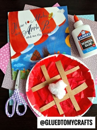 A children’s book titled How to Make an Apple Pie and See the World, a pair of scissors, a bottle of Elmers school glue, and a paper plate cherry pie craft with a cotton ball center.