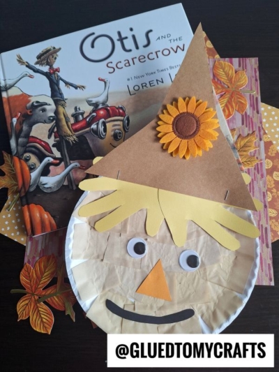 A charming paper plate scarecrow with a brown hat, yellow hair, googly eyes, and a sunflower sits on autumn-themed paper beside the book Otis and the Scarecrow by Loren Long.