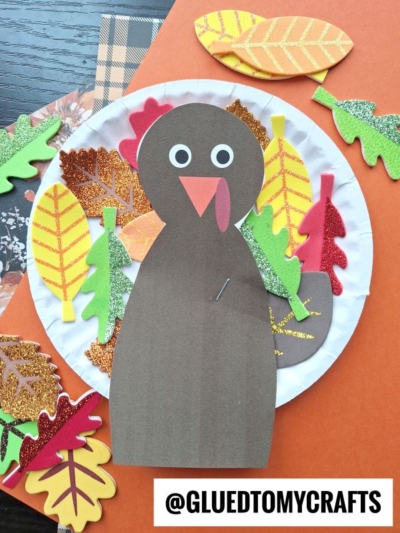 This Paper Plate Leaf Turkey craft features a brown paper turkey, surrounded by colorful glittery paper leaves. With googly eyes, an orange beak, a red wattle, and @GLUEDTOMYCRAFTS at the bottom, it’s a festive and fun Thanksgiving project!.