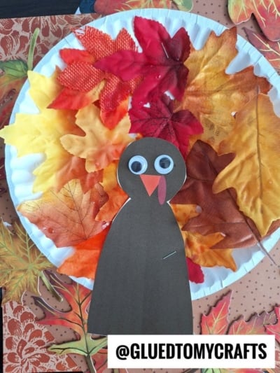 A Paper Plate Leaf Turkey craft featuring artificial autumn leaves as vibrant feathers and a cutout turkey with googly eyes, red wattle, and beak. The image includes the text @GLUEDTOMYCRAFTS at the bottom.