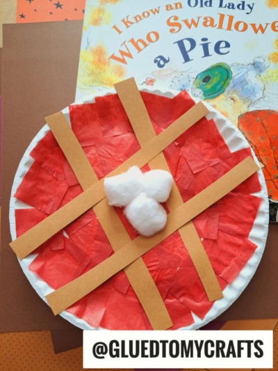 A paper plate cherry pie craft with red tissue paper, tan lattice strips, and three cotton balls in the center is shown beside the book I Know an Old Lady Who Swallowed a Pie. @GLUEDTOMYCRAFTS is labeled below.