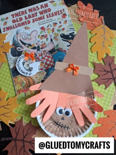 A fall-themed scene with a book, paper leaves, and a charming paper plate scarecrow craft featuring a brown hat, orange bow, and orange handprint as hair. Text @GLUEDTOMYCRAFTS appears at the bottom.