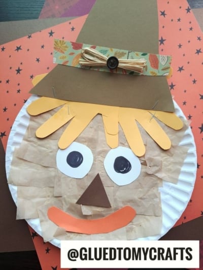 A charming paper plate scarecrow craft featuring a brown hat, yellow hair, googly eyes, a paper nose and mouth, and a bow with a button; the background showcases fall-themed paper. Text reads @GLUEDTOMYCRAFTS. This adorable plate scarecrow is perfect for celebrating the autumn season!.