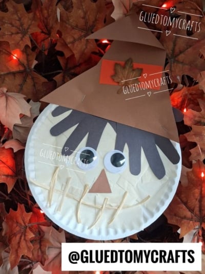 A charming paper plate scarecrow face made from a simple plate, googly eyes, a brown construction paper hat and hair, a paper nose, and a straw mouth—surrounded by artificial autumn leaves and glowing orange lights. This adorable plate scarecrow craft brings festive fall warmth to any space.