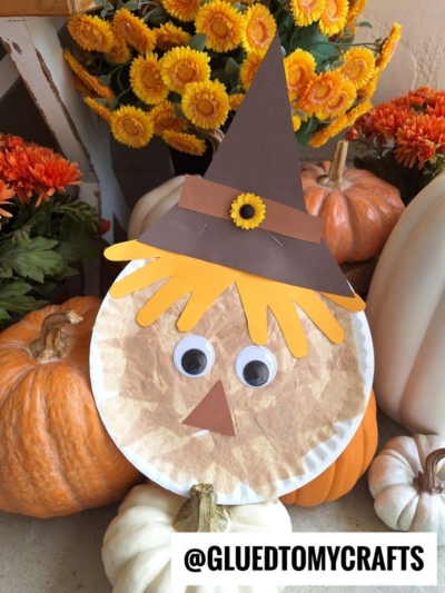 A charming paper plate scarecrow with googly eyes, a brown paper hat, and yellow paper hair is surrounded by pumpkins and orange flowers. The handle @GLUEDTOMYCRAFTS appears at the bottom.