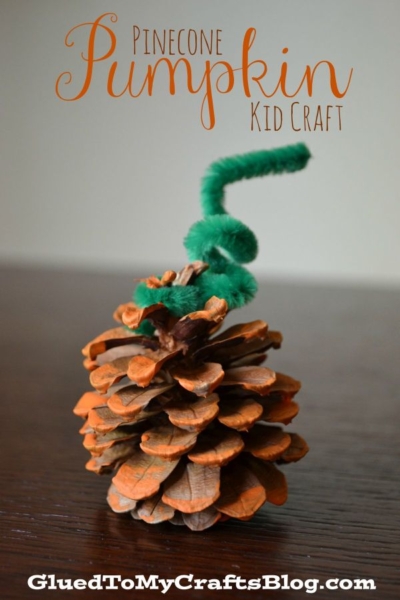 A Pinecone Pumpkin painted orange, with a green pipe cleaner twisted on top to mimic a stem, sits on a dark wooden surface. Text reads: Pinecone Pumpkin Kid Craft. GluedToMyCraftsBlog.com.