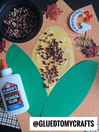 A fall-themed corn on the cob craft project perfect for preschool includes green and yellow paper shaped like corn, pinto beans glued on as kernels, an Elmer’s glue bottle, orange polka-dotted paper, faux leaves, and a bowl of pinto beans. This fun and creative activity brings the classic corn on the cob crafts for preschool to life with vibrant textures and colors.