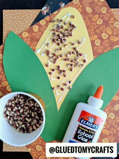 Craft project showing yellow and green paper shaped like a corn cob, with glue and seeds arranged on top—perfect for corn on the cob crafts for preschool! A bottle of Elmer’s glue and a cup of seeds are nearby, ready for little hands to use. Autumn-patterned paper serves as the background, making this corn on the cob craft project fun and festive.