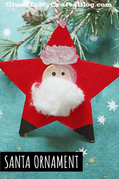 A Santa Star Ornament painted with Santa’s face, featuring a white cotton-ball beard, black eyes, and a pink nose. The red star-shaped ornament hangs in front of green pine branches on a teal background.
