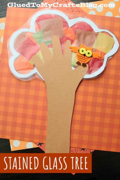 A craft project featuring stained glass fall trees made from brown paper shaped like a hand, with tissue paper leaves and a small paper owl, set against an orange checkered background.