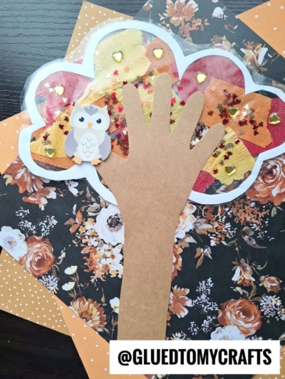 A fall craft featuring a handprint tree with an owl, inspired by Stained Glass Fall Trees, decorated with tissue paper, gold heart stickers, and glitter on a floral background. The handle @GLUEDTOMYCRAFTS is at the bottom.
