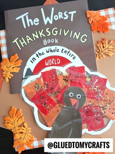 A children’s book titled “The Worst Thanksgiving Book in the Whole Entire World” is surrounded by orange paper leaves and a handmade stained glass turkey craft with a black body and colorful, textured feathers. The username @GLUEDTOMYCRAFTS appears at the bottom.