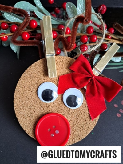 A cute reindeer ornament made from a cork circle, googly eyes, a red button nose, a bow, and clothespin antlers. Red berries and green leaves accent the background. @GLUEDTOMYCRAFTS is written at the bottom. Perfect for Cork Reindeer Ornaments!.