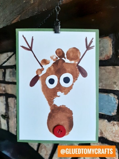 A reindeer made from brown paint thumbprints, with googly eyes, drawn antlers, and a red button nose, on white paper clipped to a green background—perfect for inspiring creative Christmas Canvas Ideas.
