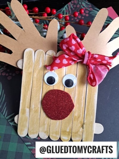 A Handprint Popsicle Stick Reindeer craft featuring popsicle sticks, cardboard antlers, googly eyes, a red glittery nose, and a heart-adorned ribbon bow. @GLUEDTOMYCRAFTS displays it among festive holiday decorations.