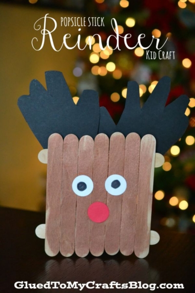 A charming Handprint Popsicle Stick Reindeer craft, handmade from brown popsicle sticks, black paper antlers, a red nose, and googly eyes, with a blurred Christmas tree in the background.