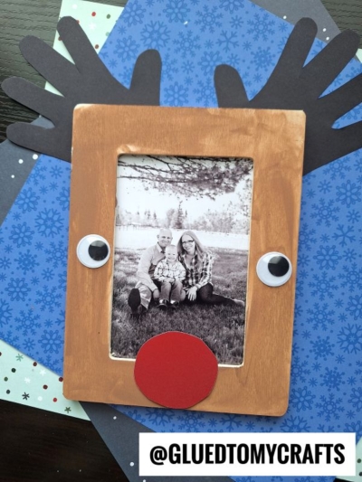 A charming Handprint Reindeer Frame with antlers, googly eyes, and a red nose displays a black and white family photo. It sits on blue snowflake-patterned paper. Handle: @GLUEDTOMYCRAFTS.