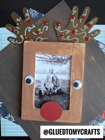 A Handprint Reindeer Frame craft with googly eyes, a red nose, and handprint antlers decorated with red gems. A black-and-white photo of two people is in the center, with @GLUEDTOMYCRAFTS at the bottom.