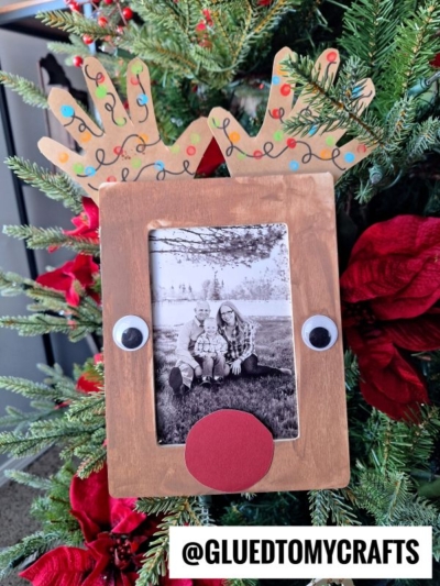 A handmade Handprint Reindeer Frame with googly eyes, a red nose, and antler cutouts holds a black-and-white photo of two people. Hanging on a Christmas tree with red poinsettias. Text reads @GLUEDTOMYCRAFTS.