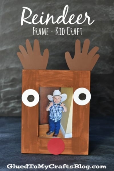 A handmade Handprint Reindeer Frame painted brown features cute handprint antlers, white eyes, and a red nose, perfectly surrounding a photo of a smiling child wearing a cowboy hat. Reindeer Frame - Kid Craft is written above.