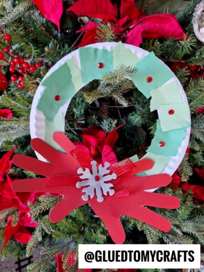 A handmade Paper Plate Christmas Wreath crafted from green paper pieces, red sequins, and paper handprints, finished with a silver snowflake and bow. This festive paper christmas wreath hangs on the tree. Text: @GLUEDTOMYCRAFTS.