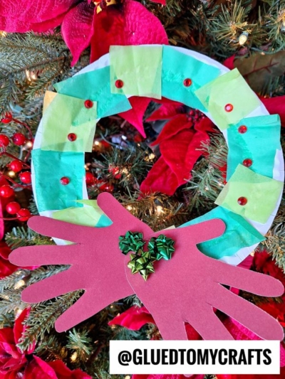 A handmade paper christmas wreath ornament with green tissue squares, red sequins, and two red paper handprints at the bottom, decorated with green bows. It hangs on a Christmas tree surrounded by festive red poinsettias.