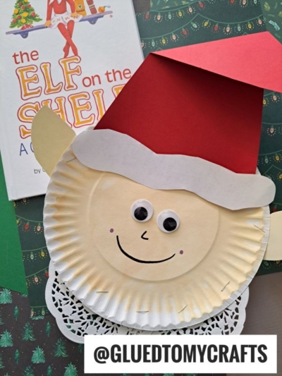A paper plate Santa craft with a red construction paper hat, white beard, googly eyes, and a smiling face sits on Christmas-themed paper next to an Elf on the Shelf book—perfect to pair with a Paper Plate Elf project. Text: @GLUEDTOMYCRAFTS.