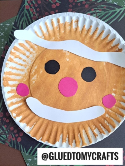 A paper plate gingerbread man painted brown is decorated with white and pink paper features to create a smiling gingerbread face, set on festive paper. @GLUETOMYCRAFTS text is at the bottom.