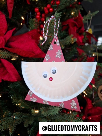 A handmade Paper Plate Santa ornament, created as a festive paper santa craft, hangs on a Christmas tree. With googly eyes, a red nose, and surrounded by red poinsettia flowers. @GLUEDTOMYCRAFTS is at the bottom.
