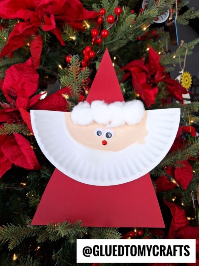 A handmade Paper Plate Santa craft made from a red triangle, a paper plate face, cotton ball hat trim and beard, and googly eyes, displayed on a decorated Christmas tree. @GLUEDTOMYCRAFTS is at the bottom.