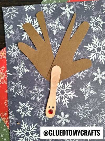 A Scoop Stick Reindeer craft made with brown handprint antlers, a wooden popsicle stick face, googly eyes, and a red nose on a snowflake-patterned background. Perfect for your next reindeer kid craft! @GLUEDTOMYCRAFTS is written below.