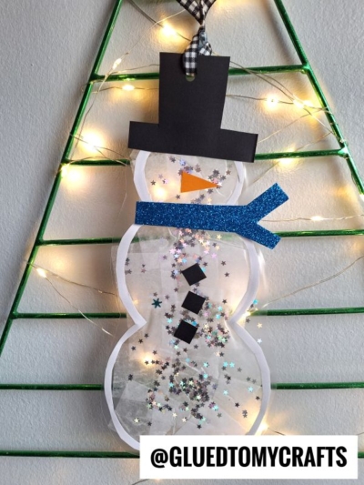 A stained glass snowman craft with a black hat, blue scarf, and star-shaped glitter inside hangs on a green triangle frame with string lights. @GLUEDTOMYCRAFTS is written at the bottom.