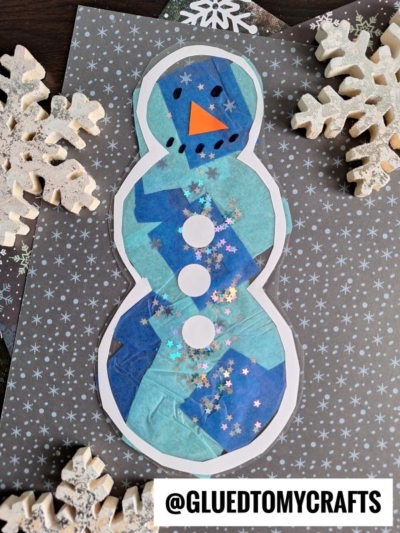 A paper snowman craft inspired by Stained Glass Snowmen, decorated with blue tissue paper, sparkly star confetti, three white buttons, and an orange triangle nose on a gray snowflake background with foam snowflakes.