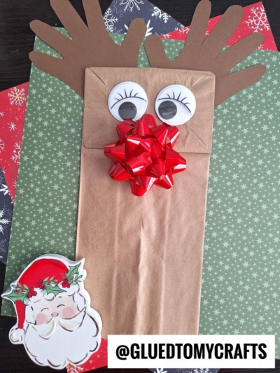 A Paper Bag Reindeer puppet decorated with googly eyes, brown handprint antlers, and a red bow nose sits on festive paper featuring a cartoon Santa and the text @GLUEDTOMYCRAFTS.